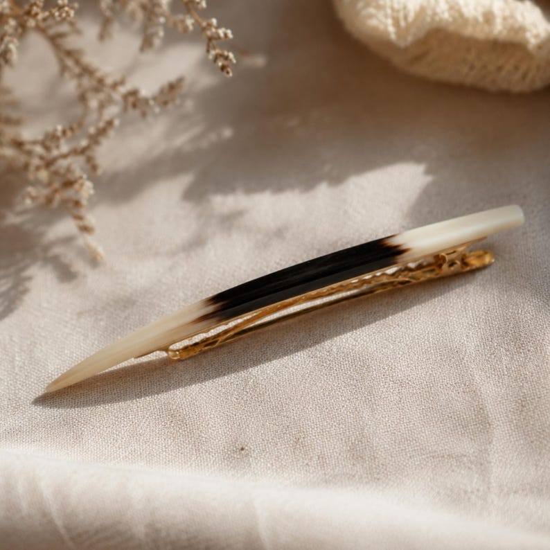 Puede incluir: Una horquilla para el cabello con una p&uacute;a larga y c&oacute;nica que presenta un degradado suave desde el color crema en la punta hasta un centro negro oscuro y de vuelta al crema en la base, sujeta a un mecanismo de pinza de cocodrilo de metal dorado.