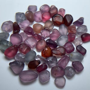 Puede incluir: Una colección de piedras preciosas pulidas en tonos rosa, morado y gris. Las piedras varían en tamaño y forma, con una apariencia suave y pulida. Las piedras preciosas están dispersas sobre una superficie blanca.