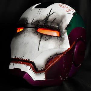 Op de afbeelding: Een op maat geschilderde helm met een witte basis, zwarte accenten en rood oplichtende ooglenzen. Het ontwerp bevat paarse, groene en rode elementen, met het woord "HA" op de bovenkant gekrabbeld. De helm heeft een verweerde, gestikte uitstraling.