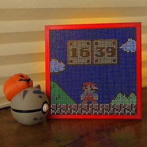 Könnte beinhalten: Ein rot gerahmtes Pixel-Art-Licht mit einer klassischen Videospielszene mit einer Figur und Landschaft. Neben dem Licht befinden sich ein grauer und weißer Pokéball und eine orangefarbene Kürbisdekoration.