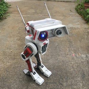 Op de afbeelding: Een gedetailleerd, wit en rood robotmodel met een rechthoekige kop, twee antennes en een blauw oplichtend oog. De robot heeft gelede benen en voeten en staat op een betonnen oppervlak.