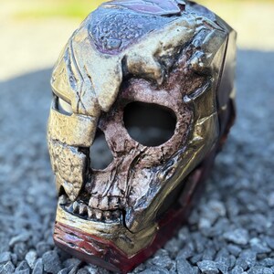 Op de afbeelding: Een gedetailleerd, verweerd masker met een schedelachtig ontwerp. Het masker heeft een combinatie van goud, bruin en rode tinten, met zichtbare texturen en ingewikkelde details. De oogkassen en mond zijn open en de algehele esthetiek suggereert een fantasie- of cosplay-thema.