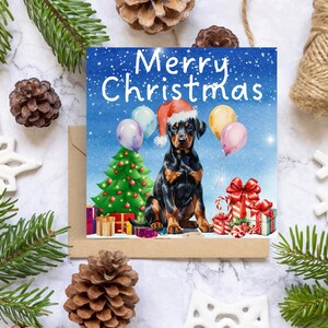 Può includere: Biglietto di Natale con un'illustrazione di un Dobermann con un cappello di Babbo Natale. La carta presenta un albero di Natale, palloncini e regali confezionati. Il testo "Merry Christmas" è scritto in alto.