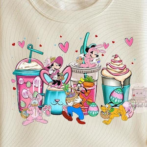 Peut inclure: Sweat-shirt crème avec un graphisme coloré sur le thème de Pâques. Le motif comprend des personnages de dessins animés, des œufs de Pâques et des boissons. Le graphisme présente des accents roses, bleus et verts, avec des cœurs dispersés.