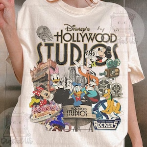 Può includere: T-shirt color crema con una grafica colorata di personaggi Disney e la scritta "Disney's Hollywood Studios". Il design include Topolino, Paperino e altri personaggi.