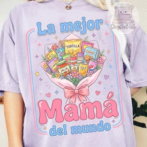 Op de afbeelding: Lichtpaars T-shirt met de Spaanse tekst "La mejor Mamá del mundo". Het ontwerp bevat een boeket van voedingsmiddelen zoals salsa en koekjes. De tekst is roze en blauw.