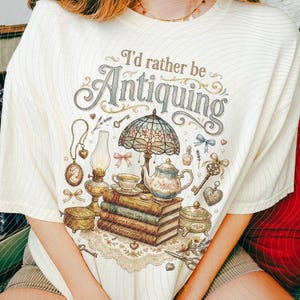 May include: Cream-colored t-shirt with the text "I'd rather be Antiquing" in a vintage font. The graphic features antique items like a lamp, teapot, books, and keys. The design is in soft pastel colors, creating a cozy, nostalgic feel. The shirt is a relaxed fit.