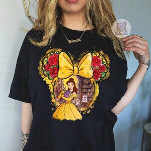 Puede incluir: Camiseta negra con un dise&ntilde;o de orejas de Mickey Mouse, con un lazo amarillo y rosas rojas. El gr&aacute;fico incluye a Bella de La Bella y la Bestia sosteniendo un libro. La palabra "Beauty" es visible en el libro.