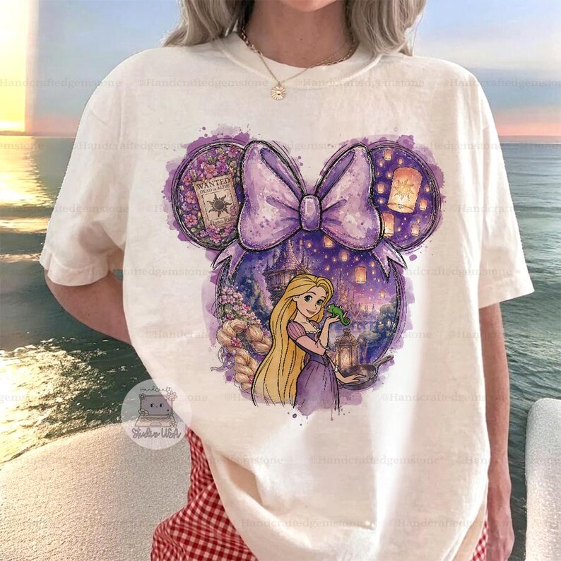 Puede incluir: Camiseta color crema con un dise&ntilde;o morado en forma de Minnie Mouse con un lazo. El dise&ntilde;o incluye una ilustraci&oacute;n de princesa, un castillo y linternas flotantes. La oreja izquierda tiene un cartel de "Wanted". Prenda de vestir informal.