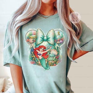 Puede incluir: Camiseta verde claro con un gr&aacute;fico colorido de Ariel de La Sirenita. El dise&ntilde;o incluye un gran lazo, conchas marinas y elementos submarinos. La camiseta es de estilo casual.