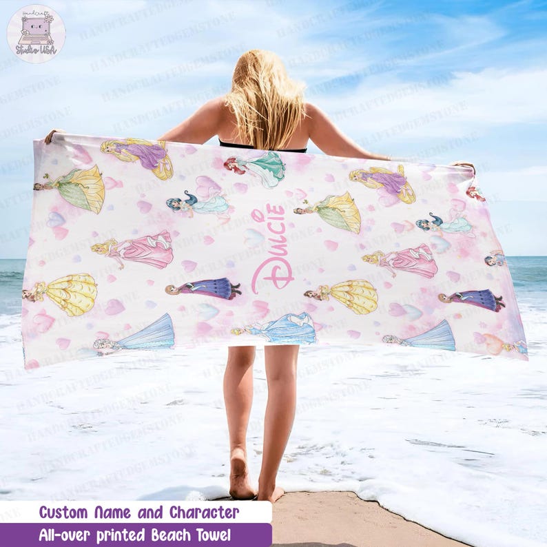 Puede incluir: Una toalla de playa rectangular con un estampado integral de varias princesas de dibujos animados con vestidos coloridos sobre un fondo blanco con corazones rosas dispersos. El nombre Dulcie est&aacute; impreso en una fuente rosa estilizada en el centro.