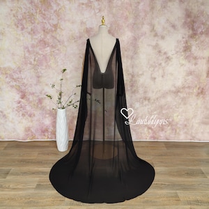 May include: A black sheer cape with a deep V-neck design, displayed on a mannequin. The cape drapes to the floor, showcasing its flowing fabric. The background features a soft, textured pink and white wall.