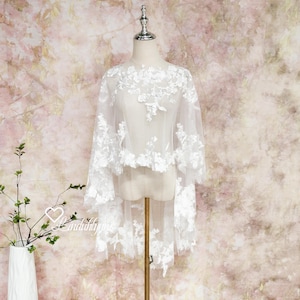 May include: A white, sheer bridal cape with floral lace detailing. The cape features a high-low hemline and is displayed on a mannequin. The delicate lace design is visible throughout the garment, creating a romantic and elegant look.
