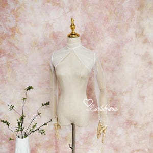 Puede incluir: Un bolero blanco transparente con mangas largas se exhibe en un maniquí. El bolero presenta un cuello alto y una tela delicada y fluida. El maniquí se encuentra sobre un fondo floral rosa y blanco. Las mangas están adornadas con pequeñas perlas blancas.
