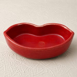 Könnte beinhalten: Eine rote Keramikschale in Form von Lippen. Die Schale hat eine glänzende Oberfläche und ein kleines, kreisförmiges Emblem am Boden. Das Innere ist tiefrot und kontrastiert mit dem etwas helleren Rand.