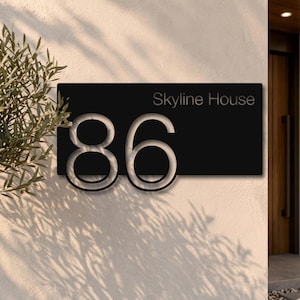 May include: A rectangular black house number sign featuring the large cutout numerals 86 and the text Skyline House in the upper right corner. The sign is mounted on a light-coloured wall, displaying a clean, modern, and high-contrast design.