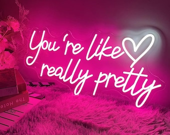 May include: A neon sign in a bright pink hue displays the phrase "You're like really pretty" in a cursive font, with a white heart symbol. The sign is placed on a fluffy pink surface.