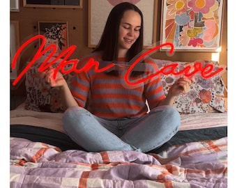 May include: A person seated on a bed holding a red neon sign that reads "Man Cave". The person is wearing a striped orange and purple sweater and blue jeans. The bed has a patterned comforter with orange and purple hues.