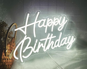 May include: A white neon sign that reads "Happy Birthday" in a cursive font. The sign is illuminated and set against a light background. A woven basket with dried flowers is in the background.