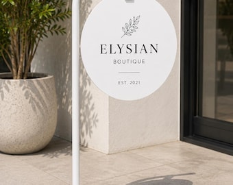 May include: A white metal floor sign stand with a circular hanging sign. The sign features a black line drawing of a leafy branch above the text ELYSIAN BOUTIQUE, a horizontal line, and the text EST. 2021. The sign is attached by a white strap.