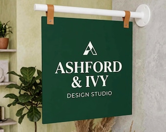 May include: A square, dark green hanging sign with white text that reads ASHFORD & IVY DESIGN STUDIO. The sign is suspended from a white horizontal rod by two tan leather straps attached to a circular white wall mount on a light gray wall.