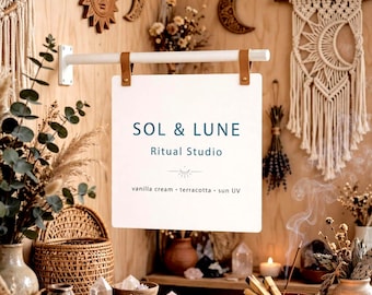 May include: A white square hanging sign with tan leather straps attached to a white horizontal rod. The sign features the blue text SOL & LUNE Ritual Studio, a small crescent moon icon, and the text vanilla cream, terracotta, and sun UV.