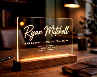 May include: A rectangular clear acrylic desk sign with warm LED illumination set in a dark wood base. The sign features the text Ryan Mitchell, Lead Counsel - Hudson Legal, Texas, and a rectangular box with the text Custom Logo Here.