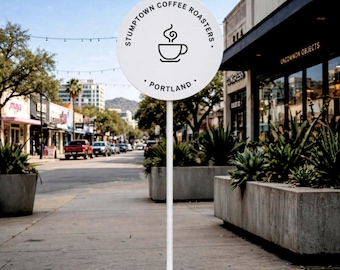 May include: A white circular sign on a thin white pole with a round base. The sign features the black text STUMPTOWN COFFEE ROASTERS and PORTLAND, with a central black line drawing of a steaming coffee cup centered on the white surface.