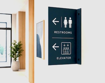 May include: A dark blue wall-mounted directional sign featuring white graphics and text. The top section shows a left arrow, restroom icons, and the word RESTROOMS. The bottom section displays a left arrow, an elevator icon, and the word ELEVATOR.