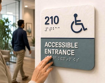 May include: A square wall sign with rounded corners featuring a white top section with the number 210 and a blue wheelchair accessibility icon, and a dark gray bottom section with the white text ACCESSIBLE ENTRANCE, both with braille.