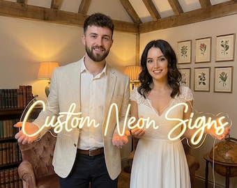 May include: A couple holds a glowing neon sign that reads "Custom Neon Signs." The sign is in a cursive font and emits a warm, inviting light. The background includes a room with wooden beams and framed botanical prints.