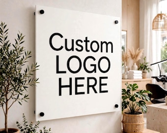 May include: A square white acrylic sign mounted on a wall with four black circular standoffs. The sign features the black text "Custom LOGO HERE" printed in a bold sans-serif font. The sign is positioned above a wooden shelf in a bright room.