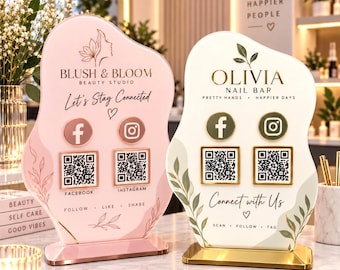 May include: Two irregular-shaped acrylic signs with gold stands. The left sign is pink with Blush & Bloom Beauty Studio branding, Facebook and Instagram icons, and QR codes. The right sign is white with Olivia Nail Bar branding and similar icons.