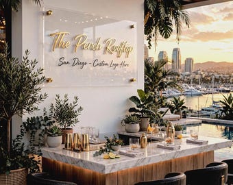 May include: A rectangular clear acrylic sign with gold lettering reading The Perch Rooftop San Diego - Custom Logo Here, mounted with four gold standoffs on a white wall above a marble-topped bar with black chairs and various barware.