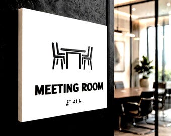 May include: A square white sign mounted on a black wall featuring a black graphic of a table and two chairs, the text "MEETING ROOM" in black capital letters, and a row of raised braille dots positioned directly below the text.