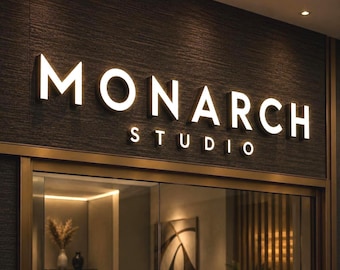 May include: A wall-mounted sign featuring the illuminated white text MONARCH STUDIO in a bold, sans-serif font. The letters are mounted on a dark, textured, horizontal-lined surface above a glass entrance with a view of an interior space.