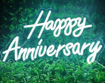 May include: A white neon sign with the words "Happy Anniversary" in a cursive font. The sign is set against a backdrop of lush, green foliage. The neon light glows brightly, creating a celebratory atmosphere.
