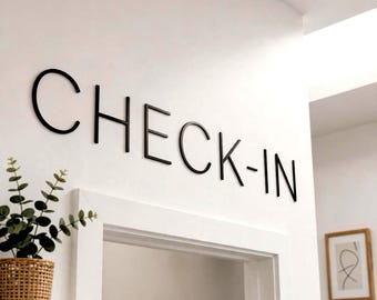 May include: A wall-mounted sign featuring the word CHECK-IN in bold, black, sans-serif capital letters. The lettering is mounted directly onto a smooth, white wall surface above a doorway, creating a high-contrast, clean, and legible display.
