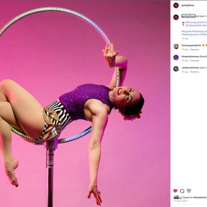 Peut inclure: Une femme v&ecirc;tue d'un haut &agrave; paillettes violet et d'un short z&eacute;br&eacute; pose sur un grand cerceau lumineux sur fond rose. Le cerceau est suspendu &agrave; un poteau, et la femme effectue une danse a&eacute;rienne. L'image est bien &eacute;clair&eacute;e et capture un moment d'athl&eacute;tisme.