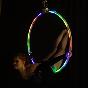 Peut inclure: Une femme en tenue noire est suspendue dans un cerceau a&eacute;rien aux couleurs de l'arc-en-ciel et brillamment &eacute;clair&eacute;. Le cerceau est illumin&eacute; par des lumi&egrave;res LED, cr&eacute;ant un affichage vibrant sur fond sombre. La femme effectue une pose acrobatique.