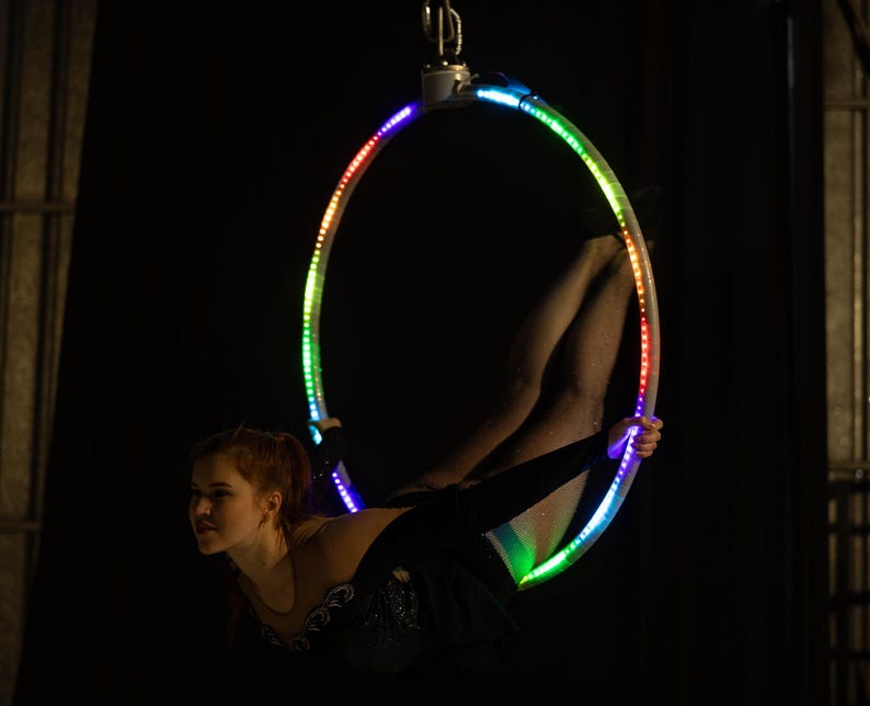 Peut inclure: Une femme en tenue noire est suspendue dans un cerceau a&eacute;rien aux couleurs de l'arc-en-ciel et brillamment &eacute;clair&eacute;. Le cerceau est illumin&eacute; par des lumi&egrave;res LED, cr&eacute;ant un affichage vibrant sur fond sombre. La femme effectue une pose acrobatique.