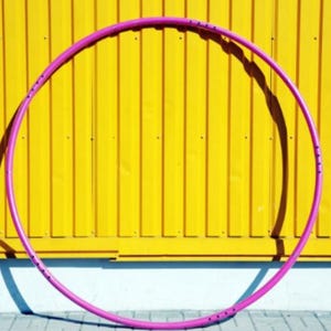 Può includere: Un hula hoop rosa acceso contro un muro di legno giallo. Il cerchio è circolare e sembra essere fatto di plastica. L'immagine è ben illuminata, con l'hula hoop che proietta un'ombra sul muro. Un articolo per il fitness o il tempo libero.