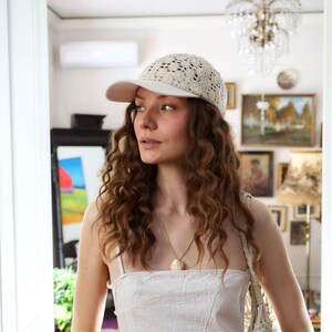 May include: A cream-colored baseball cap with a lace-like design on the crown and a solid brim. The cap is worn by a person with long, curly brown hair. A necklace with a shell pendant is also visible.