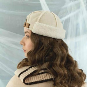May include: A cream-colored, fleece-lined hat with a brown leather strap and buckle. The hat has a rounded crown and a folded-up brim. The person is also wearing a cream-colored sweater with black and cream stripes.