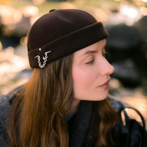 May include: A dark brown brimless cap featuring a rolled edge and a silver-toned metal chain detail attached to the side. The cap has a smooth, felt-like texture and a structured, rounded crown that sits close to the head.