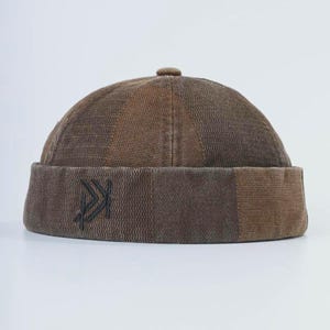 Könnte beinhalten: Braune Skullcap mit einem genähten Design. Die Kappe hat ein dunkles, gesticktes Logo auf der Vorderseite. Der Hut ist aus einem strukturierten Stoff gefertigt und hat eine runde Krone. Die Farbpalette umfasst Braun- und Beigetöne.