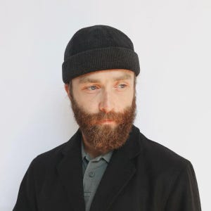 Puede incluir: Un gorro de pana negro con un borde doblado. El gorro lo lleva una persona con barba completa y una chaqueta negra. El fondo es una pared blanca lisa. El gorro es de estilo clásico.