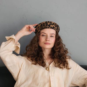 May include: A leopard print beret with a black and brown pattern. The beret is worn by a person wearing a light beige button-down shirt. The background is a solid gray wall.