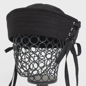 Op de afbeelding: Een zwarte docker-pet met een brede, gestikte rand en een vetersluiting aan de zijkant met zilverkleurige metalen oogjes. De pet heeft lange, platte zwarte stoffen linten die aan de zijkant hangen, gemaakt van een stevig, effen textielmateriaal.