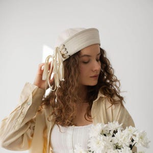 May include: A cream-coloured hat with a flat top and a decorative band. The hat features a series of eyelets on the side, with cream-coloured fabric strips hanging down. The hat is worn by a person with curly brown hair.